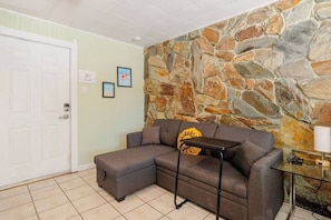 A living room with a sectional sofa, a stone accent wall, a door, and a small table with a lamp. - Quiet Studio with Sunny Private Yard (Wilton Manors)