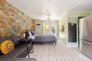 A bedroom with a stone wall, a bed, a gray sofa with a sun pillow, a small table, a ceiling fan, and a view through a glass door. - Quiet Studio with Sunny Private Yard (Wilton Manors)