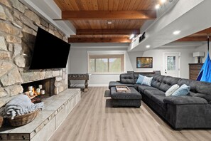 A living room with a stone fireplace, a flat-screen TV mounted on the wall, a sectional sofa, and a wooden ceiling. - Peaks View A-Frame: Hot Tub, Trails, A/C, andEV Charger (Flagstaff)