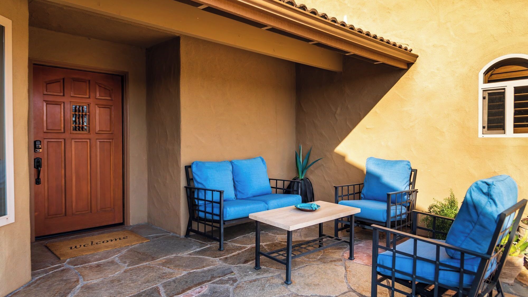 A patio with a stone floor, a wooden door, and blue cushioned chairs and a table.