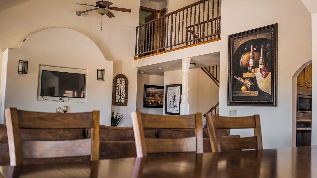 A dining area with wooden chairs and a table, a ceiling fan, a TV mounted on the wall, and a framed artwork depicting still life.