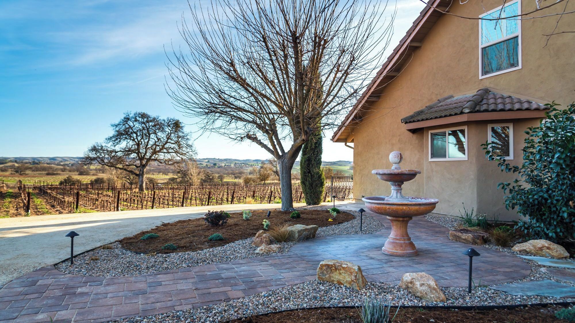 A house with a vineyard in the background, a stone pathway, and a decorative fountain.