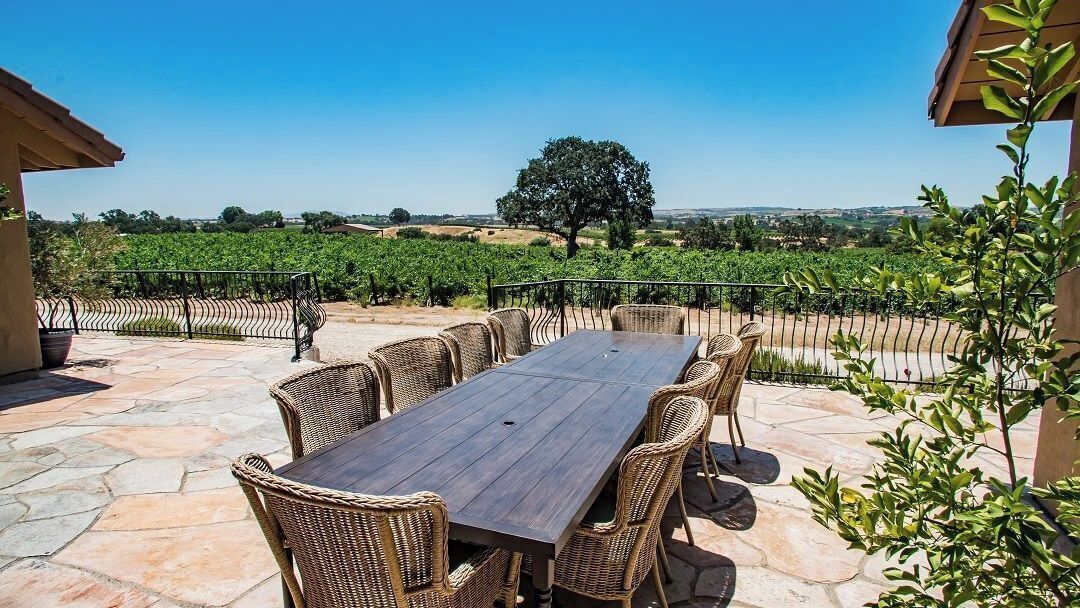 An outdoor patio with a long table and wicker chairs, surrounded by stone flooring and a metal fence.