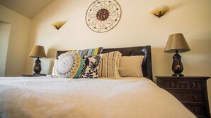 Una cama bien hecha con una colcha y almohadones estampados, flanqueada por dos mesitas de noche con lámparas, en una habitación con un adorno decorativo sobre la cama.
