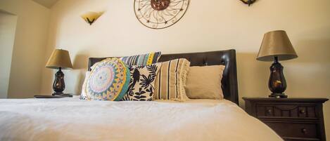 A neatly made bed with a patterned blanket and pillows, flanked by two bedside tables with lamps, in a room with a decorative wall piece above the bed.
