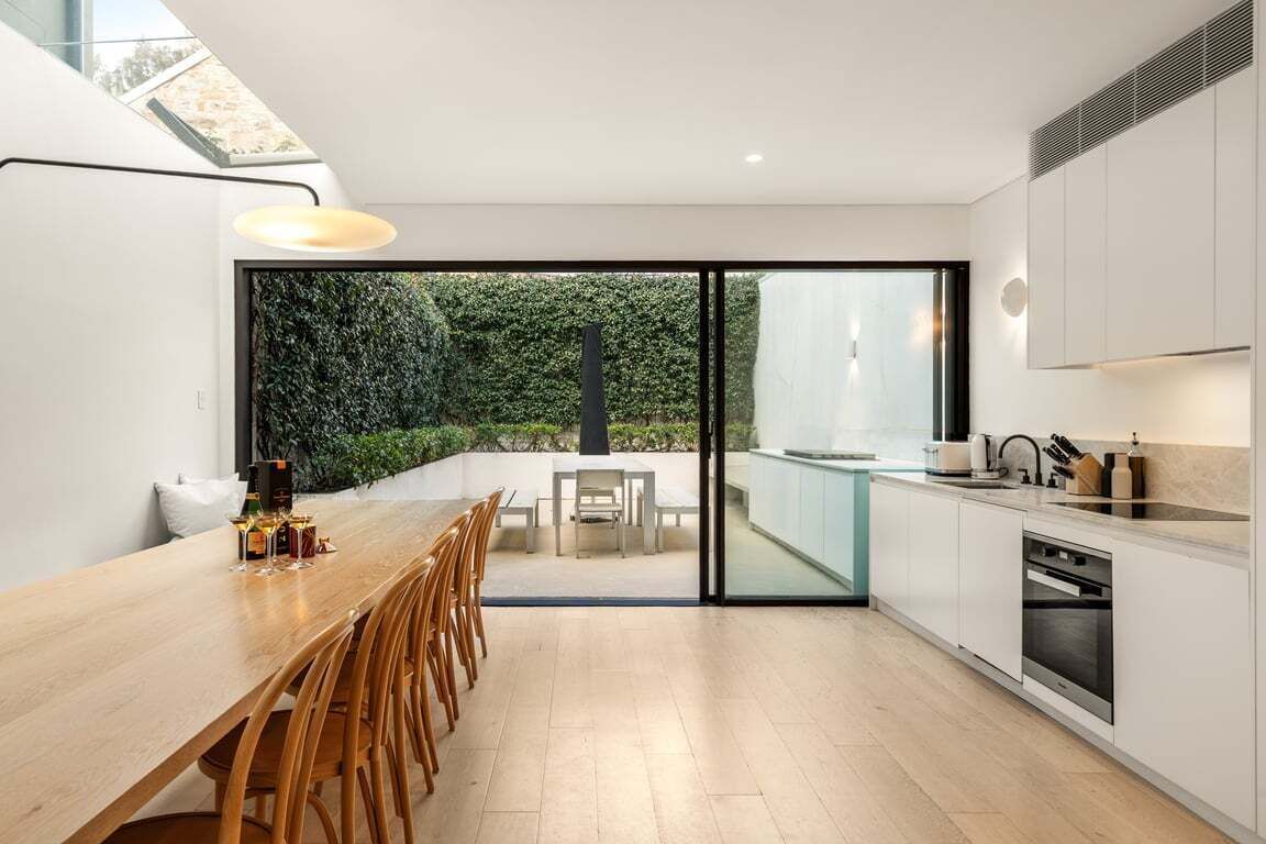A modern kitchen with a dining area, large windows, and a view of greenery.