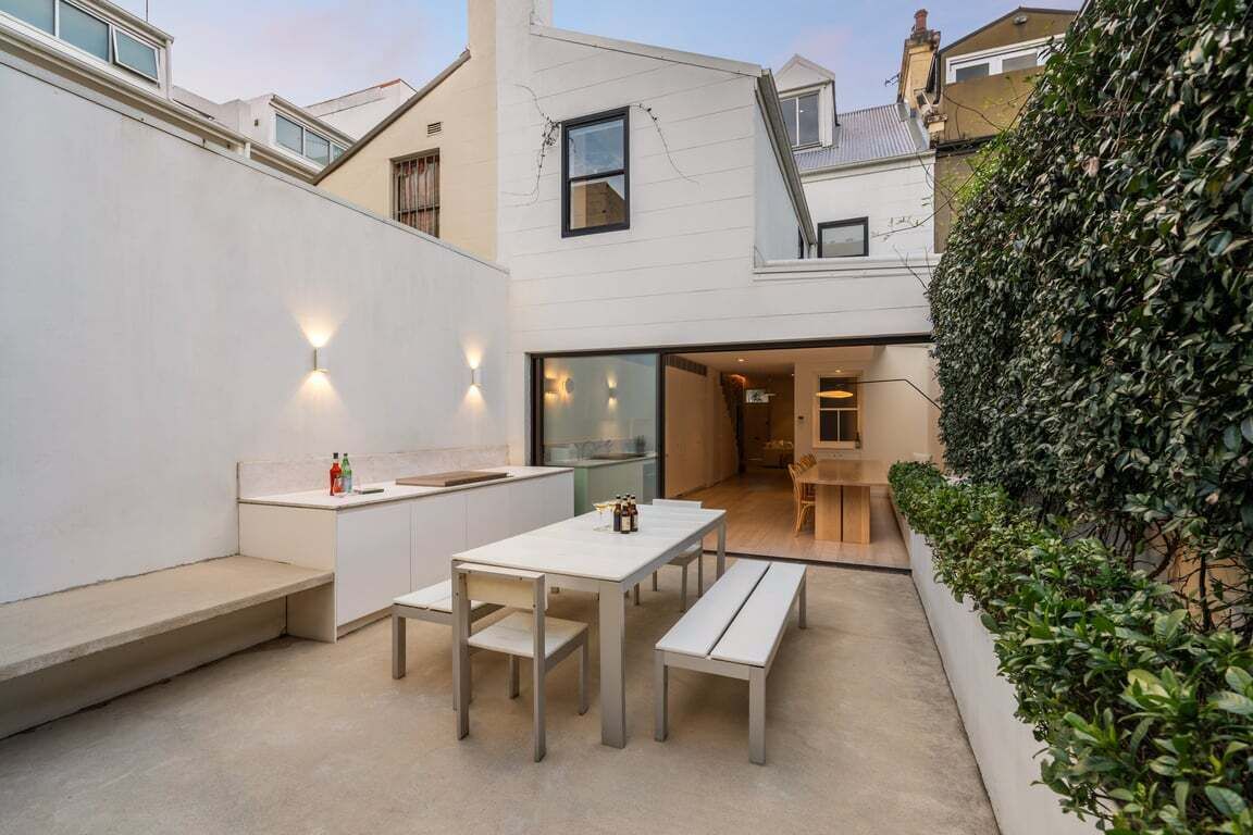 Un área de comedor exterior moderna con banco y mesa, una pequeña cocina con mostrador, y un seto a la derecha.