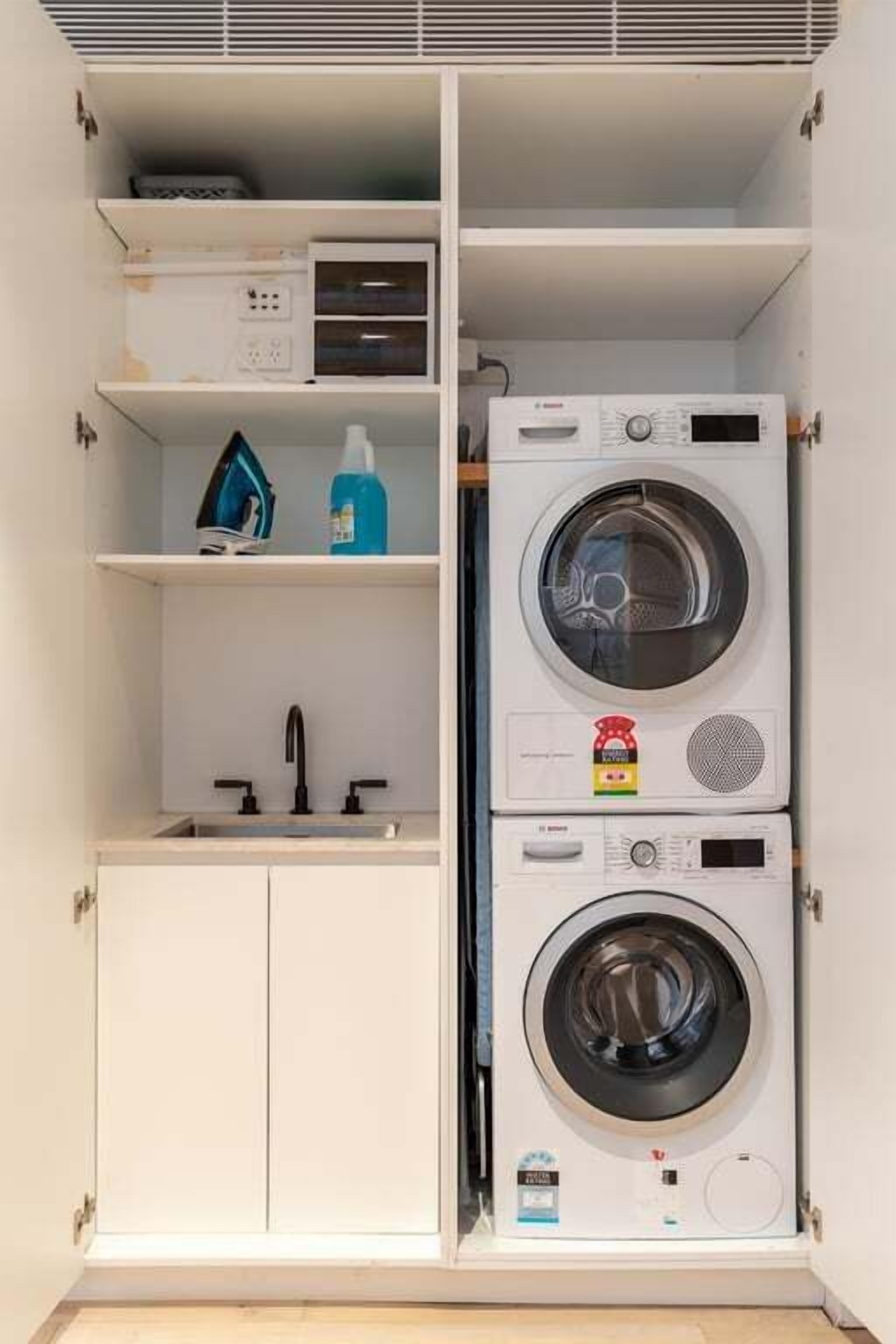 A laundry room with two stacked washing machines, a shelf with cleaning supplies, and a sink.