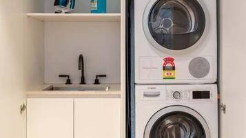 A laundry room with two stacked washing machines, a shelf with cleaning supplies, and a sink.