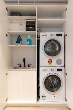 A laundry room with two stacked washing machines, a shelf with cleaning supplies, and a sink. - Palmer Darlinghurst - Stunning Terrace Home (Darlinghurst)