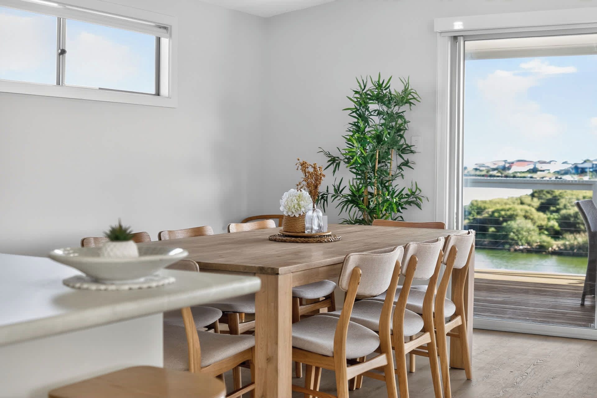 Comedor con mesa y sillas de madera, un jarrón con flores y vistas a zonas verdes y edificios a través de una ventana.