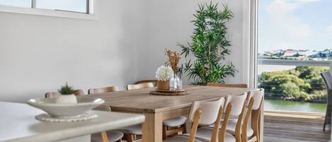 Comedor con mesa y sillas de madera, un jarrón con flores y vistas a zonas verdes y edificios a través de una ventana.
