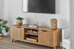 A wooden console with a flat-screen TV mounted above it, a woven basket, a small framed picture, and a woven lantern on the console. - Marina Breeze - Excelsior Pde (Hindmarsh Island)