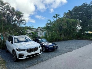 Zwei geparkte Autos, ein weißer SUV und ein schwarzer Sportwagen, auf einem Schotterweg mit einem Haus und üppiger Vegetation im Hintergrund.