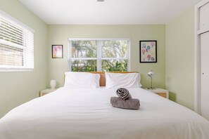 A hotel room with a bed, two bedside tables, a window with blinds, and two framed pictures on the wall. - Sunny Apartment in Quiet Lush Neighborhood (Wilton Manors)
