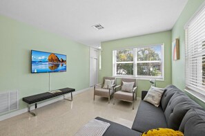 A living room with a television, seating area, and large windows. - Sunny Apartment in Quiet Lush Neighborhood (Wilton Manors)