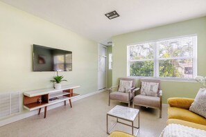 A living room with a flat-screen TV, a white console table, two armchairs, a yellow sofa, and a window with blinds. - Sunny Apartment in Quiet Lush Neighborhood (Wilton Manors)