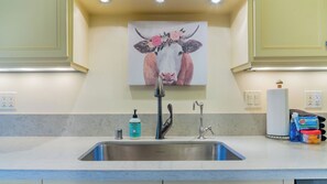 A kitchen with a stainless steel sink, a faucet, a soap dispenser, and a decorative painting of a cow on the wall. - Vin Rouge 51 by Avantstay Sleeps 8 + Hillside Deck + Ping Pong 15-min Walk to Downtown (Paso Robles)