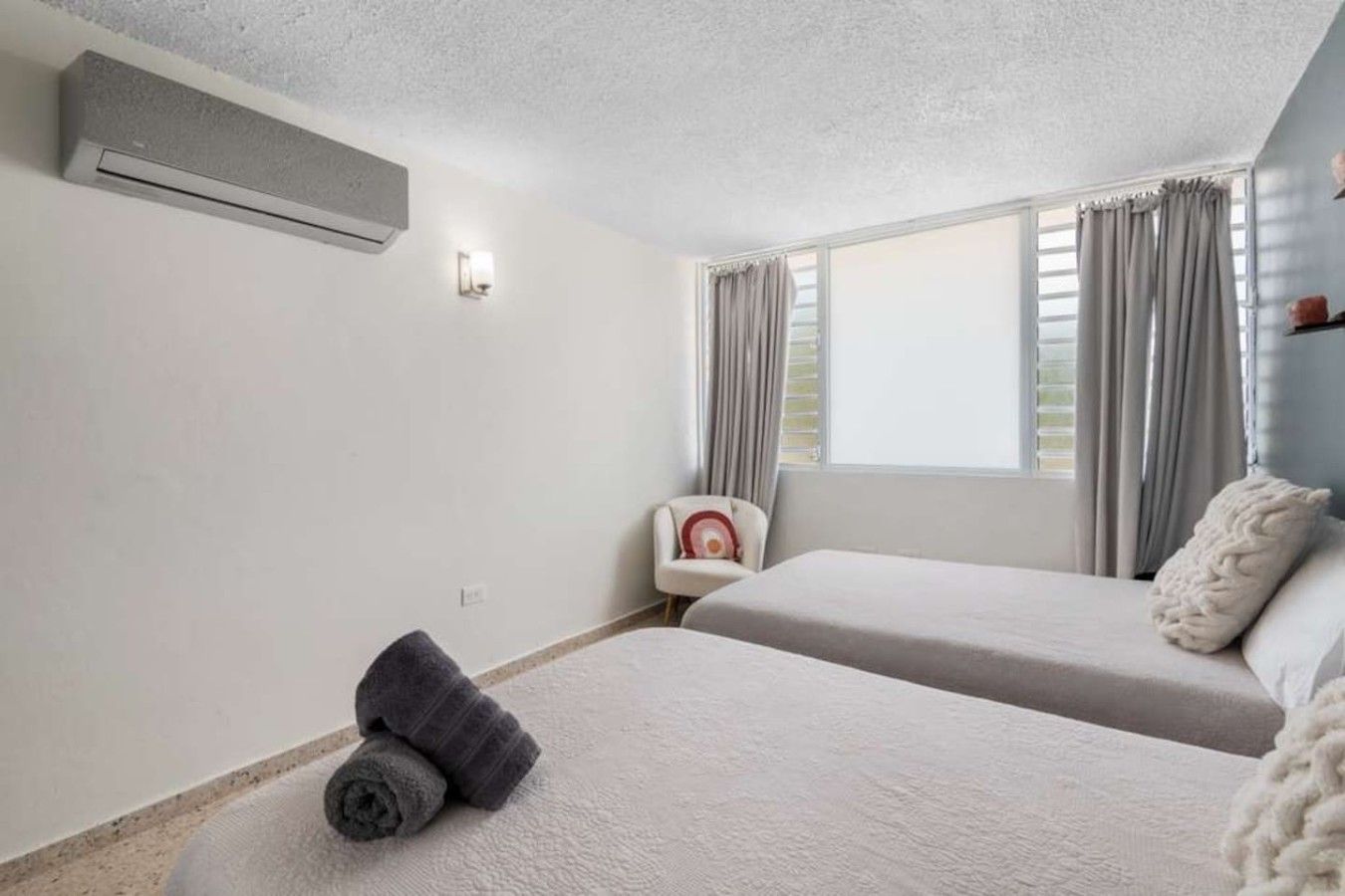 A room with two beds, a chair, a window with curtains, and an air conditioning unit.