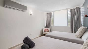 A room with two beds, a chair, a window with curtains, and an air conditioning unit.