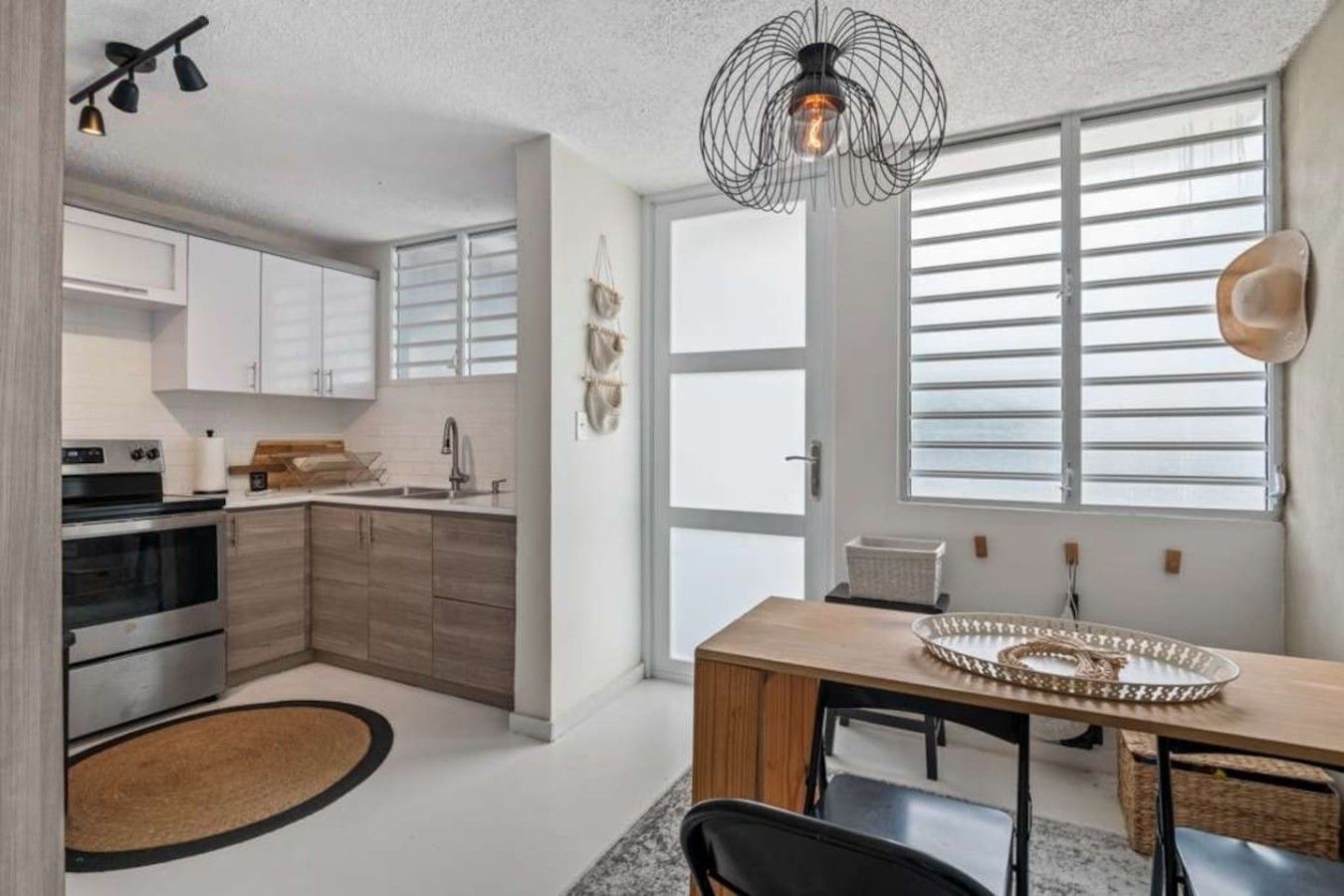 A modern kitchen with a dining area, featuring a large window, a hanging light fixture, and a central island.
