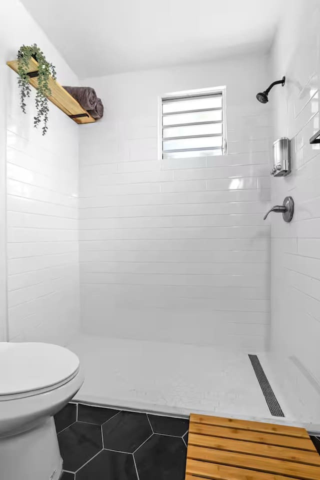 A bathroom with white tiled walls, a black hexagonal floor, a wooden bench, a toilet, and a small window.