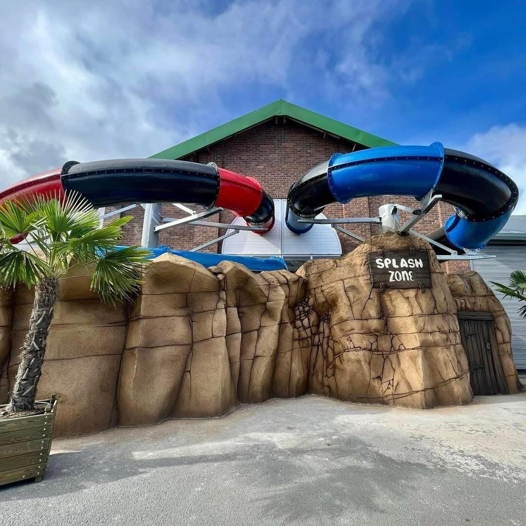 L’entrée d’un parc aquatique, avec une pancarte indiquant « Zone de jeux aquatiques » et deux toboggans.