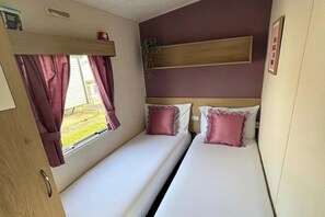 A compact room with two beds, a window with curtains, a shelf above the beds, and a small plant. - Coastal Caravan at Lyons Robin Hood, Rhyl (Rhyl)