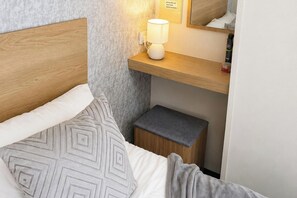Ein ordentlich bezogenes Bett mit einem gemusterten Kopfkissen, ein Nachttisch mit einer Lampe und ein kleiner Hocker.