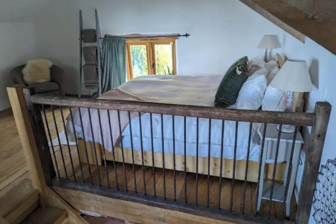 A bedroom with a wooden bed, a ladder, a chair, a desk, and a window with curtains.
