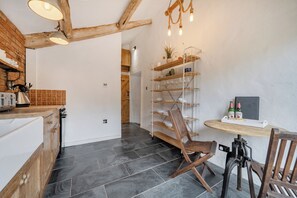 A kitchen with a wooden counter, a kettle, and a coffee machine. There is a wooden chair and a table with a bottle of beer.
