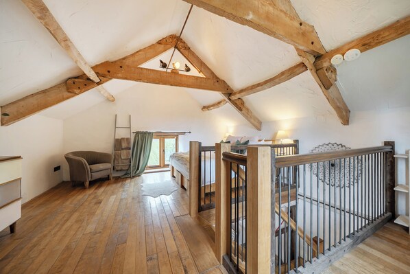 A loft bedroom with exposed wooden beams, a wooden staircase, a sofa, and a view of the outdoors.