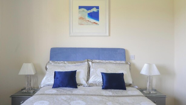Ein ordentlich bezogenes Bett mit blauem Kopfteil, zwei Kissen und einer weißen Decke. Auf jeder Seite des Bettes stehen jeweils eine Nachttischlampe.