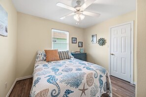 A bedroom with a bed, a ceiling fan, a window with blinds, a door, and wall decorations. - The Love Shack, 3-bedroom house in serene Galveston with AC for relaxing stays, (Galveston)
