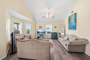 A spacious living room with a flat-screen TV, two sofas, a ceiling fan, and a large painting. - The Love Shack, 3-bedroom house in serene Galveston with AC for relaxing stays, (Galveston)