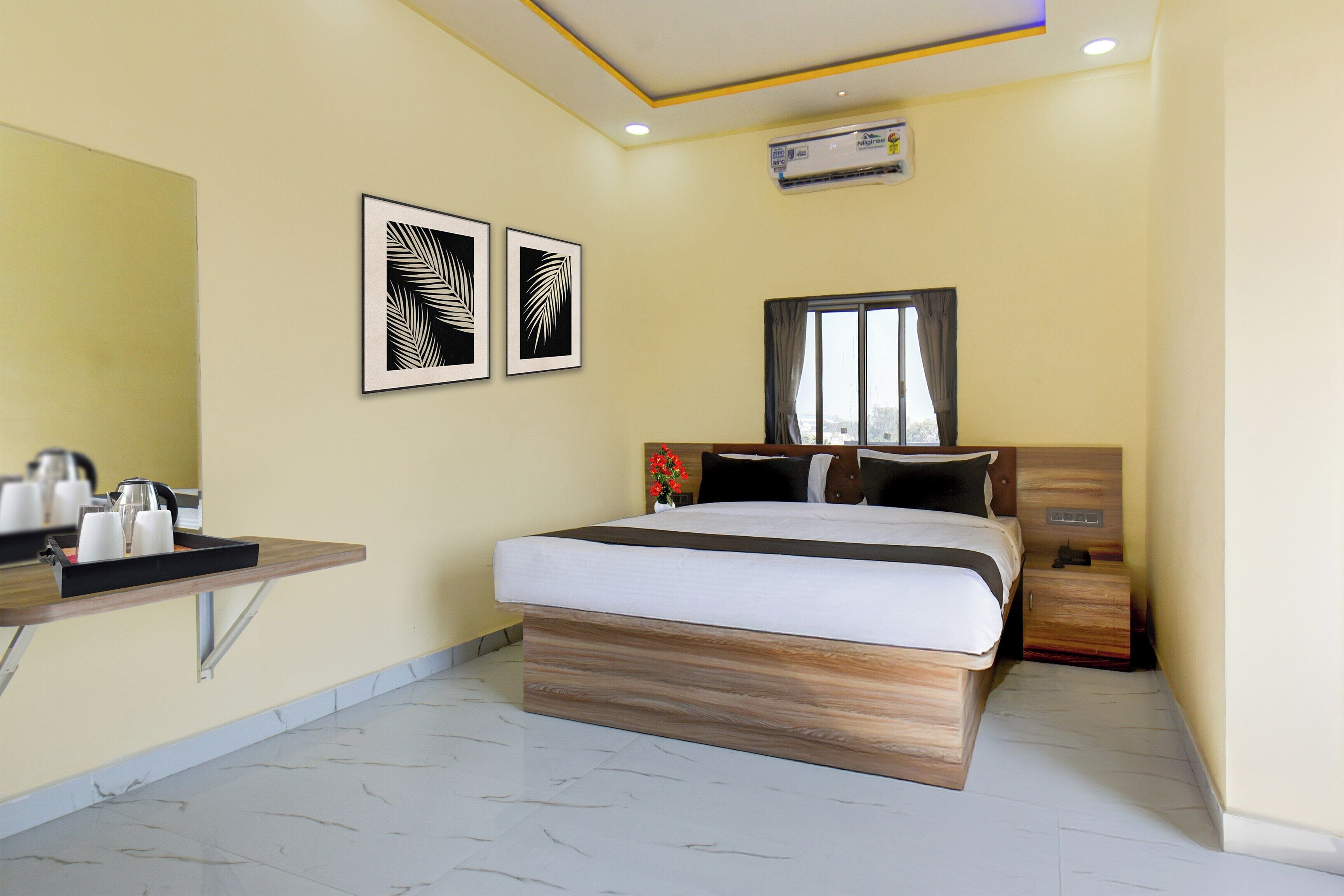 A hotel room with a bed, bedside table, a small desk with a kettle and cups, a window with curtains, and two framed leaf patterns on the wall.