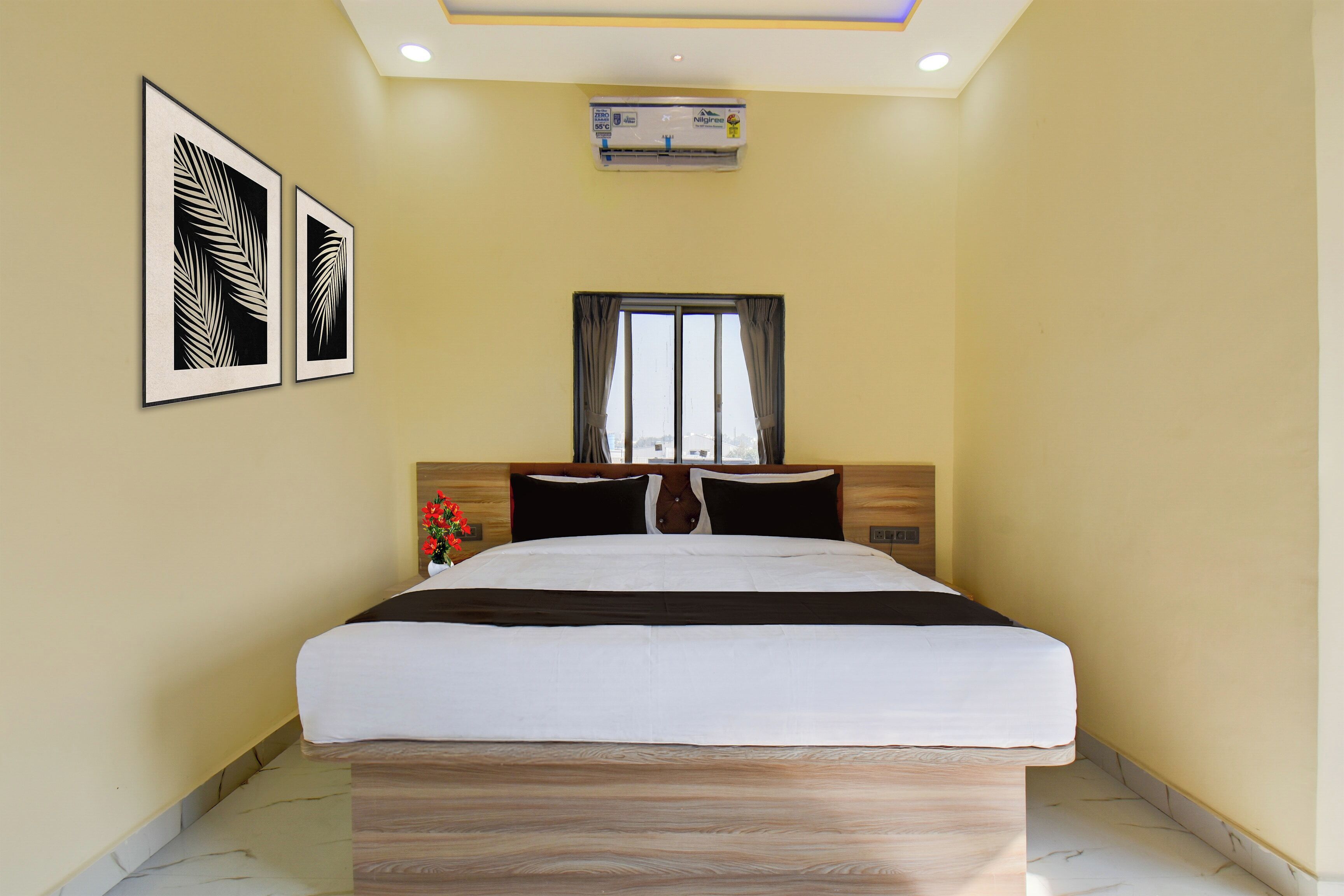A hotel room with a bed, a window with curtains, a wall-mounted air conditioner, and two framed pictures.