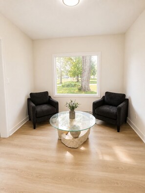 Una habitación con una mesa redonda de vidrio, dos sillas negras, una ventana con vista a árboles y paredes de tonos claros.