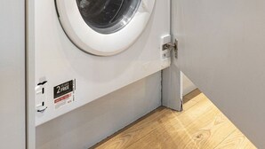 A front-loading washing machine with a digital control panel, a 2-year warranty sticker, and a red item on top. - GuestReady - Charming Loft in Vibrant London (London)