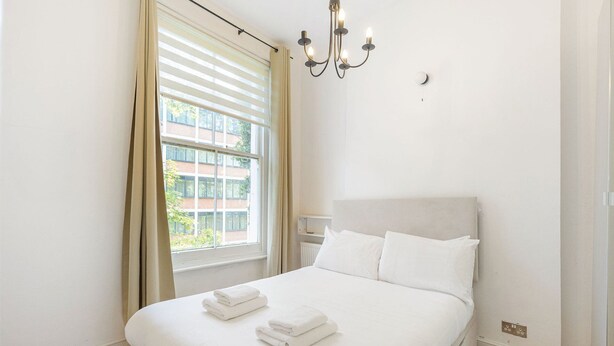 A hotel room with a bed, a window with blinds, a chandelier, and a radiator. - GuestReady - Charming Loft in Vibrant London (London)