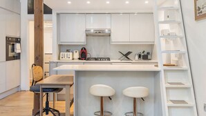 A compact kitchen with white cabinets, a gas stove, a toaster, and a coffee maker. There is a wooden chair and a small table with two bar stools. - GuestReady - Charming Loft in Vibrant London (London)