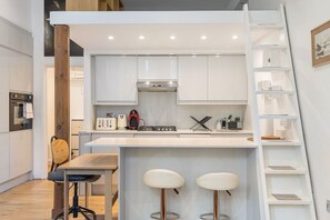 Eine kompakte Küche mit weißen Schränken, einem Gasherd, einem Toaster und einer Kaffeemaschine. Ein Holzstuhl und ein kleiner Tisch mit zwei Barhocker sind ebenfalls vorhanden.