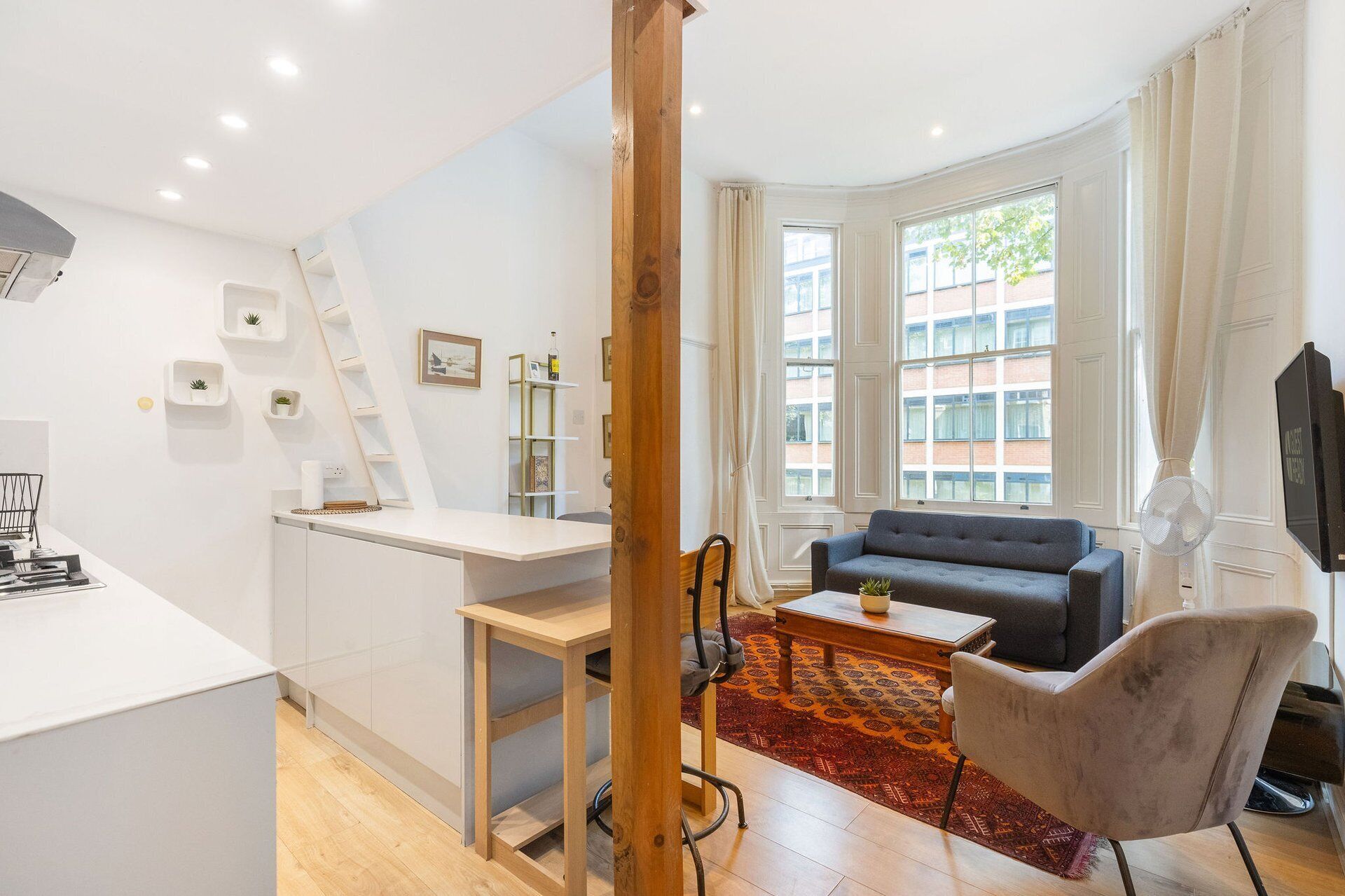 Una sala de estar moderna con un sofá, una mesa de centro y un sillón. Hay un escritorio con una silla y una escalera. Una ventana deja entrar luz natural y ofrece vistas a edificios exteriores.