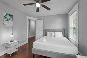 Un dormitorio con cama, mesita de noche, ventilador de techo, puerta y ventana.