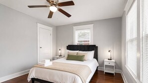 A bedroom with a bed, bedside tables, a ceiling fan, a window with blinds, and a door.