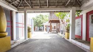 Un passage couvert mène à un bâtiment à la façade rouge et blanche, doté d’un escalier et d’un petit espace extérieur agrémenté de plantes.