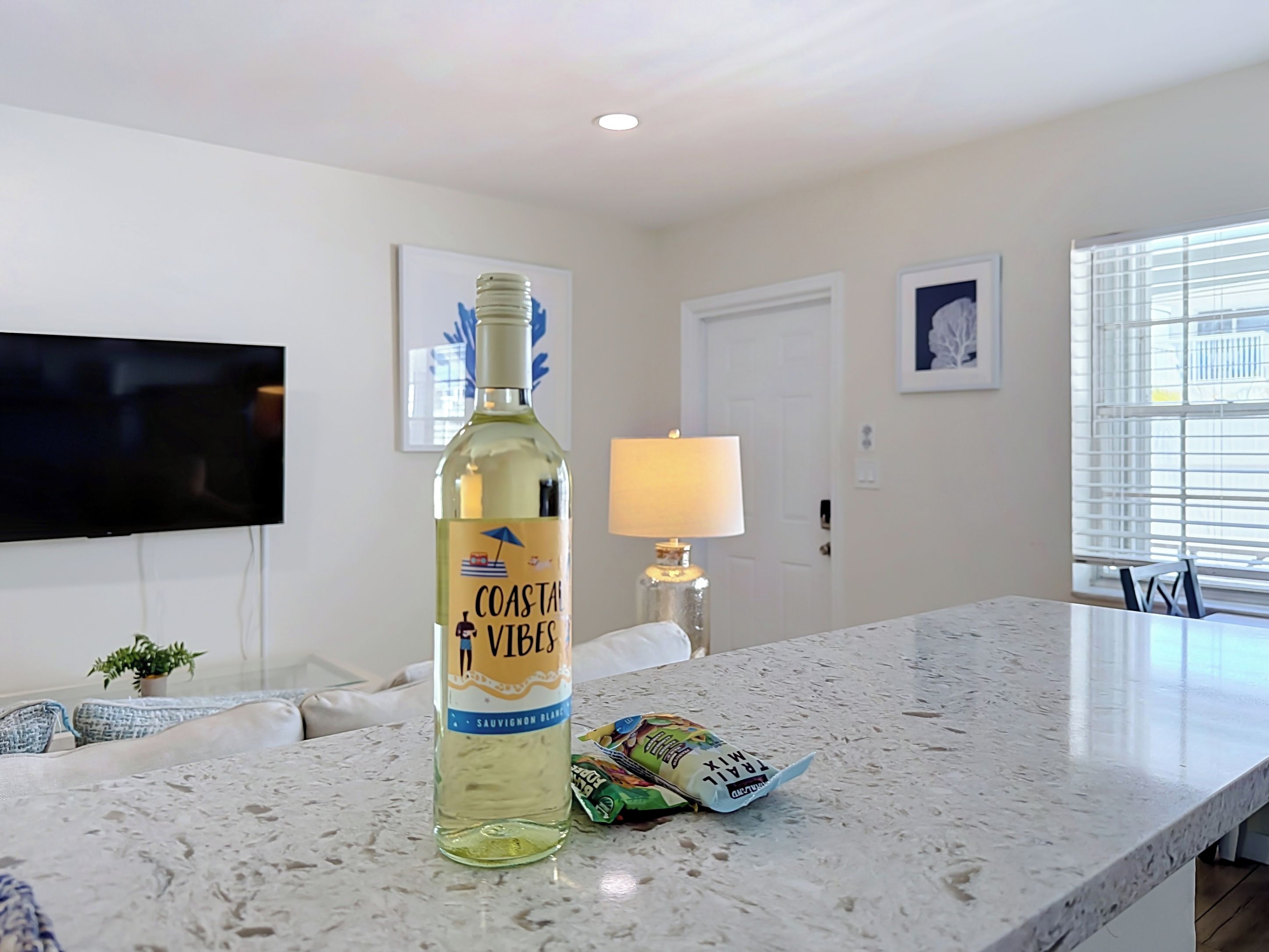 Eine Flasche Coastal Vibes Sauvignon Blanc auf einer Küchenarbeitsplatte mit Fernseher, Lampe und Snacks.