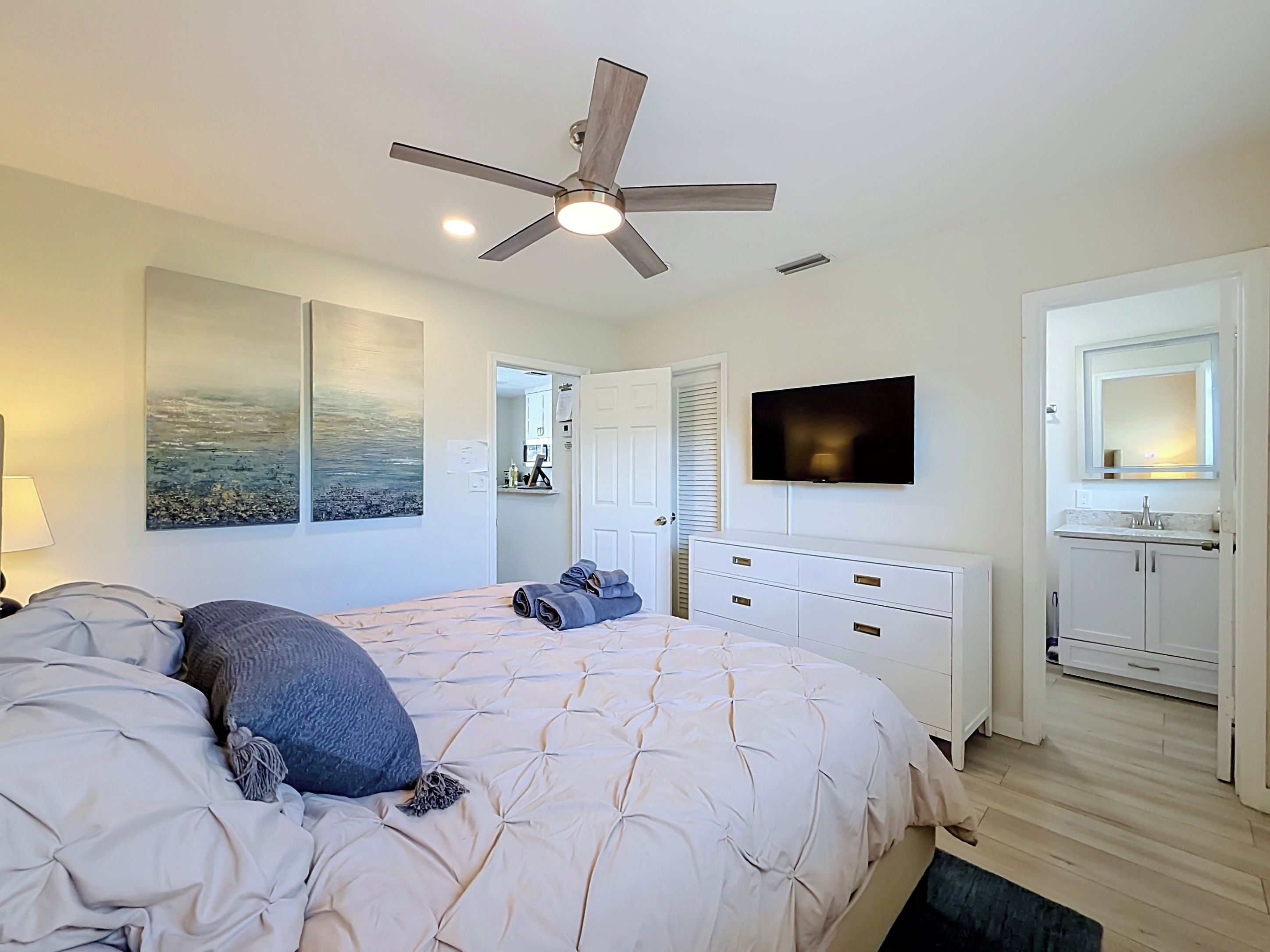 Un dormitorio con cama, ventilador de techo, televisor, baño con espejo y una obra de arte en la pared.
