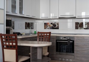 A modern kitchen with white cabinets, a built-in oven, and a dining area with a round table and wooden chairs. - Tauron Arena Cosy Apartment (Kraków)