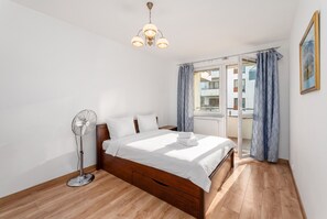 A bedroom with a wooden floor, a bed with white bedding, a standing fan, and a window with curtains.